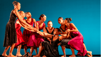 Eight dancers performing at Philadanco in red dress costumes and a blue stage background. They are all holding each other, reaching for one dancer on the floor